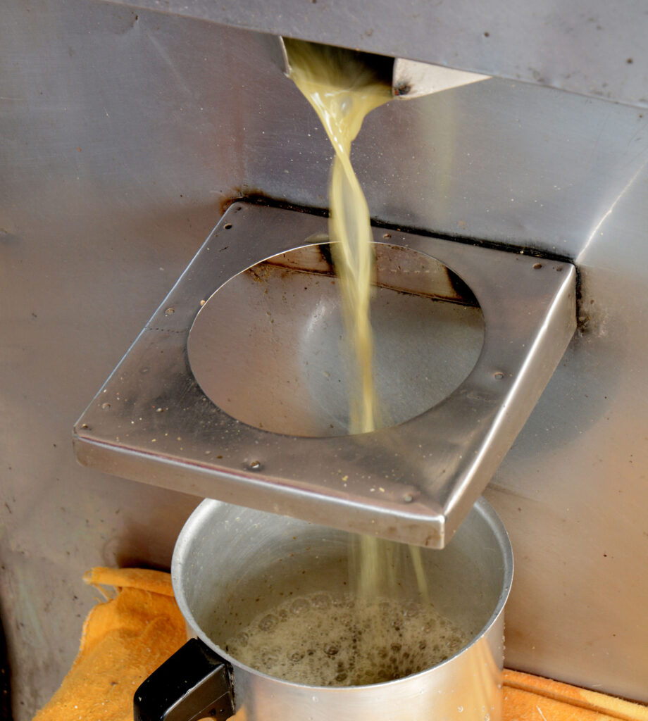 Fotografia de um moedor de cana de açúcar. Na imagem temos o caldo de cana saindo da máquina e caindo em uma panela de alumínio.