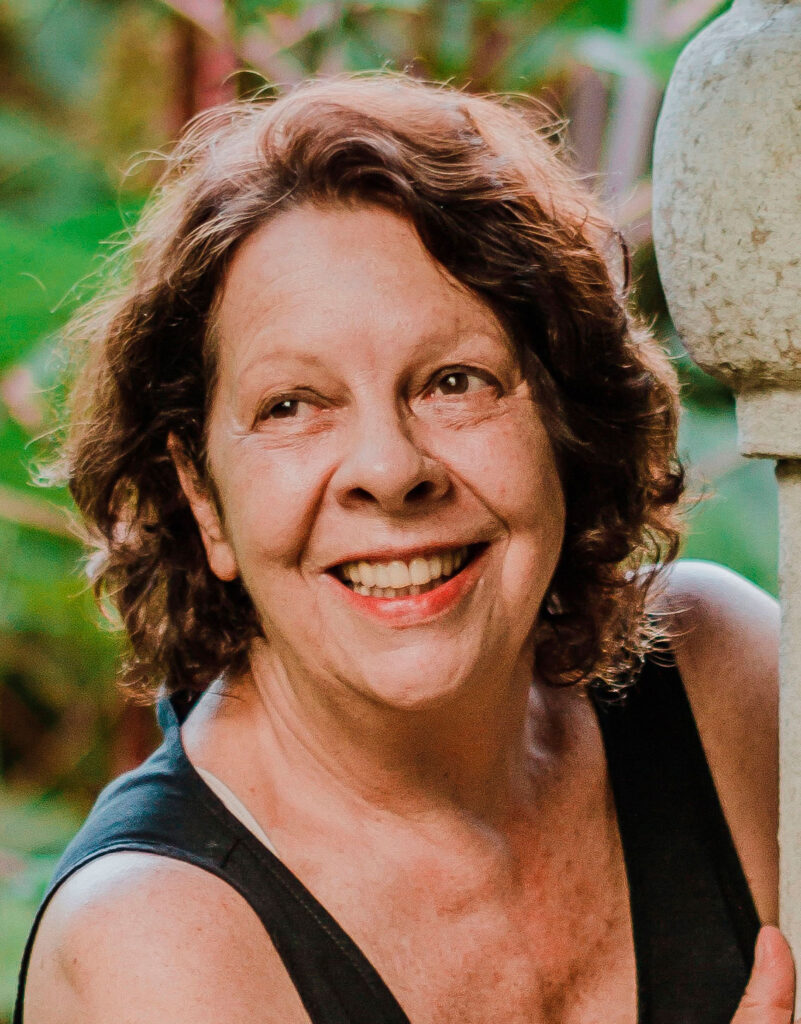 Lorane Barreto posa para foto, em uma paisagem repleta de plantas. Ela está encostada em uma estrutura de cerâmica e olha para a frente sorrindo.